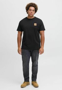 Schwarzes Baumwoll-T-Shirt mit orangefarbenem rechteckigem Logo auf der linken Brust, kombiniert mit dunkelgrauen Jeans und hellbraunen Stiefeln.