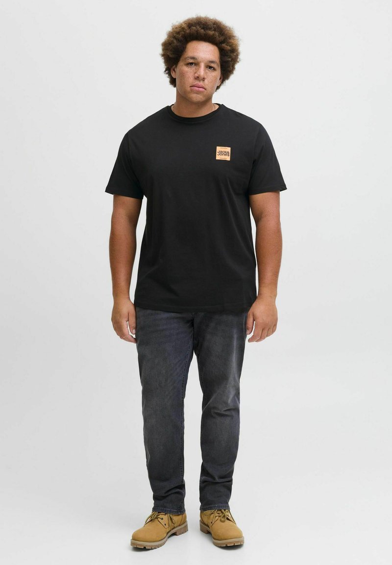 Schwarzes Baumwoll-T-Shirt mit orangefarbenem rechteckigem Logo auf der linken Brust, kombiniert mit dunkelgrauen Jeans und hellbraunen Stiefeln.