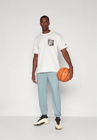 Valge lühikeste varrukatega T-särk musta graafikuga taskuga, millele on sobitatud heledad sinised dressipüksid ja musta-valge mustriga tossud, hoides korvpalli.