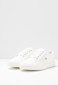 Sneakers bianchi in canvas con punta tonda, suola in gomma testurizzata e accentatura con logo dorato sul lato. Presentano lacci bianchi e dettagli cuciti.