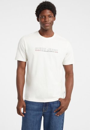 Balta medvilninė marškinėliai su pilku ir raudonu "GUESS JEANS" tekstu. Trumpi rankoviai ir apvali iškirptė, derinami su mėlynais džinsais.