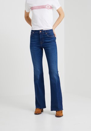 Vrouw draagt nauwsluitende donkerblauwe bootcut spijkerbroek, witte T-shirt met rode strepen en logo, en bruine loafers met hak, staand met handen achter de rug.