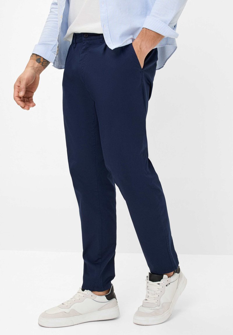 Marineblaue, schmal zulaufende Hose mit glatter Textur, versehen mit seitlichen Taschen und einem Knopfverschluss, kombiniert mit weißen Sneakern und einem hellblau gestreiften Hemd.