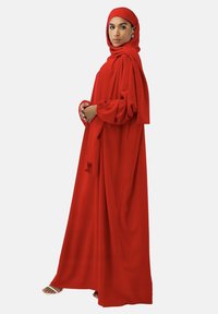 Robe rouge longue en tissu fluide, présentant une coupe ample et de longues manches. Accompagnée d'un hijab rouge assorti et de sandales.