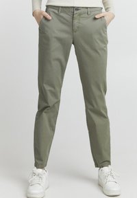 Olivově zelené chino kalhoty vyrobené z hladké látky, se bočními kapsami, slim fit a jemnými švy, v kombinaci s bílými teniskami.