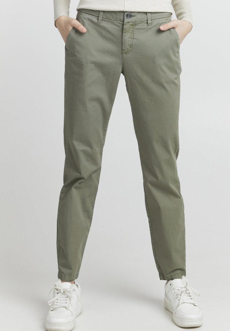 Olivově zelené chino kalhoty vyrobené z hladké látky, se bočními kapsami, slim fit a jemnými švy, v kombinaci s bílými teniskami.