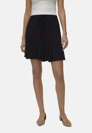 Kobieta w czarnej spódnicy za kolano z wiązaniem w talii, białej bluzce bez rękawów i beżowych butach na niskim obcasie, stojąca na jednolitym tle.