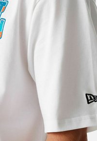Camiseta blanca de algodón con cuello redondo, que presenta un gráfico colorido en el frente y un pequeño logo negro en la manga. Textura suave.