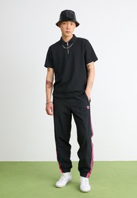 Polo shirt nera con colletto a zip, abbinata a pantaloni sportivi neri con strisce rosa ai lati. Indossata con un cappello a secchiello nero e sneakers bianche.