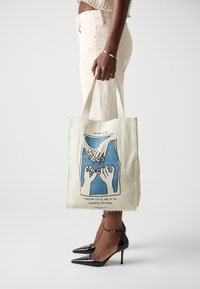 Canvas tote bag in light beige featuring a graphic design of hands and text in blue. Bag has dual handles and a sturdy structure.