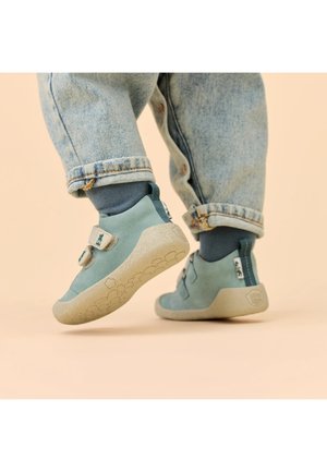 Kind draagt blauwe sneakers met klittenbandsluitingen en blauwe sokken, plaats één voet omhoog tegen een effen achtergrond.