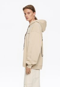 Sudadera con capucha beige de corte relajado, que presenta un cordón con herrajes decorativos y una gran impresión gráfica en la parte posterior.