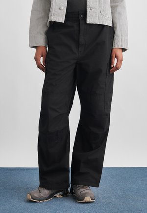 Άτομο που φοράει φαρδιά μαύρη παντελόνα τύπου cargo, γκρι ορειβατικά παπούτσια και ανοιχτόχρωμο ριγέ σακάκι, στέκεται σε μπλε χαλί.