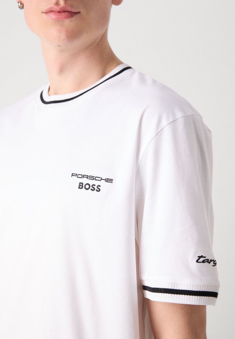 T-shirt bianca in cotone con scollo rotondo bordato di nero, presenta "PORSCHE" e "BOSS" ricamati sul petto, e "targ" sulla manica.