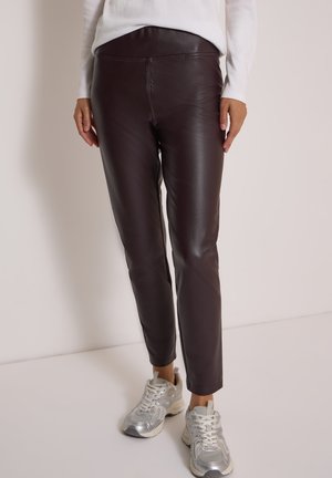 Femme portant un pantalon en cuir marron foncé et des baskets argentées, debout devant un fond clair uni.