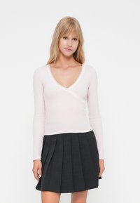 Top rosa chiaro a maniche lunghe con design a incrocio e pizzo lungo il collo. Abbinato a una gonna plissettata nera.