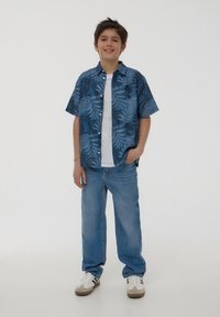 Camicia a maniche corte blu con motivo tropicale e foglie verdi, indossata sopra una maglietta bianca; abbinata a jeans azzurri chiari e sneakers bianche.