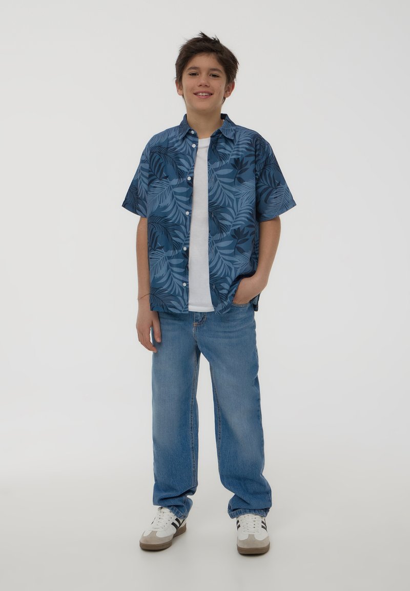 Camicia a maniche corte blu con motivo tropicale e foglie verdi, indossata sopra una maglietta bianca; abbinata a jeans azzurri chiari e sneakers bianche.