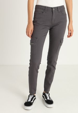 Schmal geschnittene, dunkelgraue Cargo-Hose mit vorderem Reißverschluss, seitlichen Reißverschlusstaschen, getragen mit schwarz-weißen Schnürsneakern und einem weißen, gerippten Top.