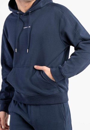 Mężczyzna w granatowej bluzie z kapturem Calvin Klein i pasujących spodniach dresowych, z gestem ręki w kieszeni, na jednolitym białym tle.