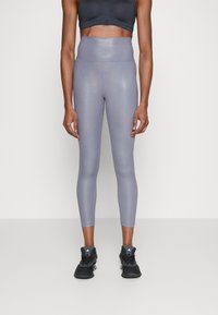 Žena oblečená v lesklých fialových legínách s vysokým pasem, černém sportovním podprsence a černých sportovních botách stojí na neutrálním pozadí.