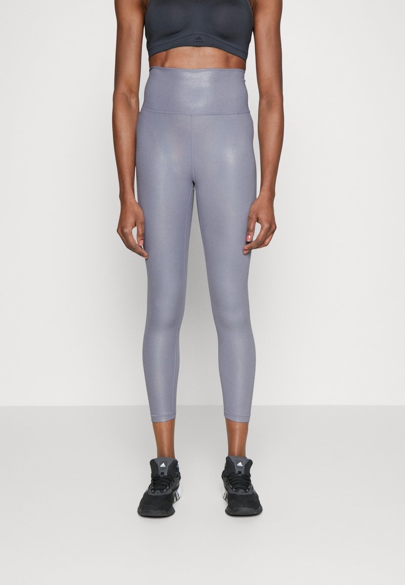 Žena oblečená v lesklých fialových legínách s vysokým pasem, černém sportovním podprsence a černých sportovních botách stojí na neutrálním pozadí.