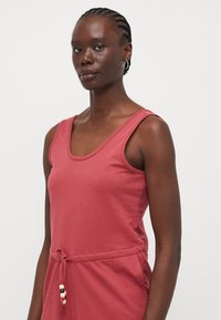 Top sans manches de couleur rose avec un décolleté rond, une taille cintrée nouée avec un cordon et des accents décoratifs en perles. Texture de tissu lisse.
