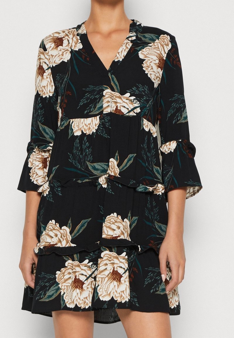 Svart blommig klänning med beige pionmönster, V-ringning, trekvartsärmar, volangskikt och en lätt tygkvalitet.