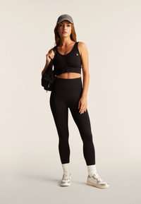 Donna che indossa un top corto nero, leggings neri, cappellino grigio, calzini bianchi e scarpe da ginnastica argento, che tiene una borsa a tracolla nera.