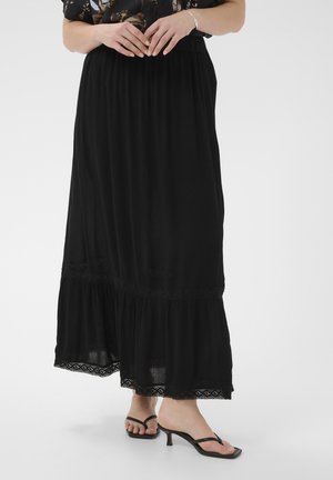 Frau trägt einen schwarzen bodenlangen Rock mit Spitzensaum und schwarze Sandaletten mit Absatz, Hände vor der Taille verschränkt.