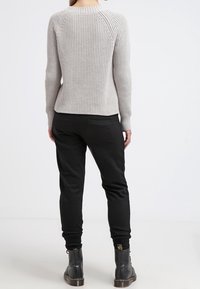 Frau steht mit dem Rücken zu uns, trägt einen hellgrauen gerippten Pullover, schwarze Hosen und schwarze Schnürstiefel vor einem weißen Hintergrund.