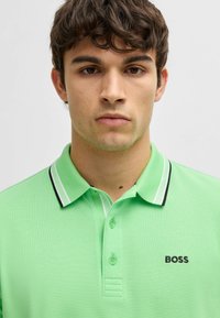 Grön polo skjorta med textilstruktur, tvåknappsknäppning och kontrasterande svart och vit randig krage. Logotypen säger "BOSS" i svart.