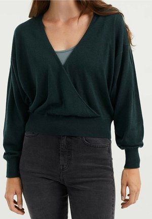 Woman wearing dark green wrap-style long sleeve sweater over a grey ribbed top paired with black high-waisted jeans.