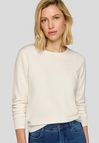 Hellbeiger Pullover mit langen Ärmeln und rundem Ausschnitt. Weiche Textur und gerippte Bündchen. Kombiniert mit blauen Denim-Jeans.