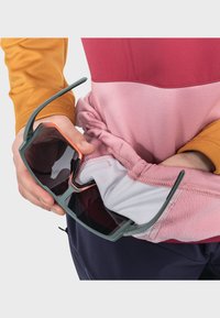 Sportsonnenbrille mit einem grauen Rahmen und dunklen Gläsern, gehalten von einer Person, die ein farblich abgestimmtes Oberteil in Pink und Orange trägt. Sichtbare gerippte Textur.