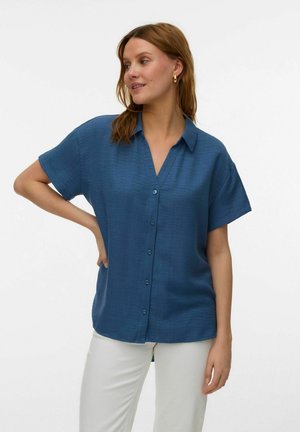 Kvinne med skulderlangt hår iført blå kortermet skjorte med knapper og hvite bukser, står med én hånd på hoften.