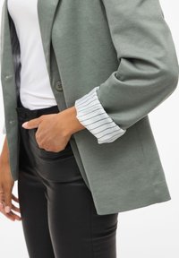 Lichtgroene blazer van textuurstof, met twee knopen en opgerolde mouwen die een wit gestreept shirt eronder onthullen, gecombineerd met zwarte broek.