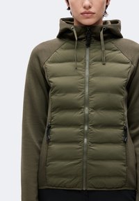 Personne portant une veste matelassée vert olive avec capuche, fermeture éclair complète à l’avant et poches latérales zippées, montrée du cou aux hanches.