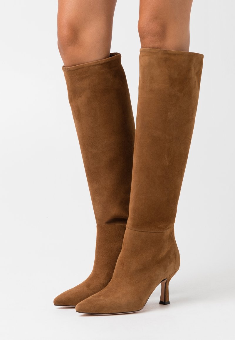 Braune Wildleder-Stiefel bis zum Knie mit spitzen Zehen und dünnen, mittelhohen Absätzen, getragen an nackten Beinen, vor einem weißen Hintergrund gezeigt.