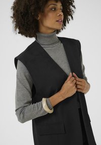 Black sleeveless vest with a smooth texture and side pockets, worn over a gray turtleneck sweater. A chunky beige bangle adorns one wrist.