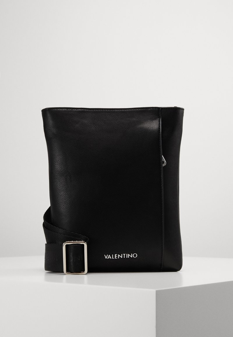 Černá kožená kabelka přes rameno s hladkým povrchem, obdélníkovým tvarem a stříbrnou sponou na nastavitelném popruhu. Obsahuje vypouklé jméno značky.