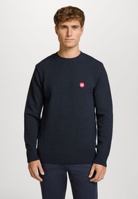 Blå strikket genser med ribbestrikket hem og mansjetter. Har en liten rød logo på brystet og rund hals. Myk tekstur.