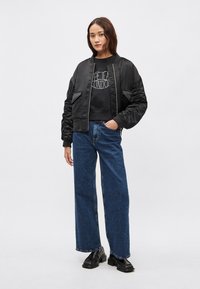 Čierna bomber bunda s rebrovaným golierom a manžetami, kombinovaná s čiernou mikinou s potlačou a širokými modrými džínsami. Čierne topánky dopĺňajú vzhľad.