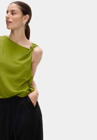 Haut vert asymétrique avec un détail de épaule noué, fabriqué en tissu léger. Associé à un pantalon noir ample.