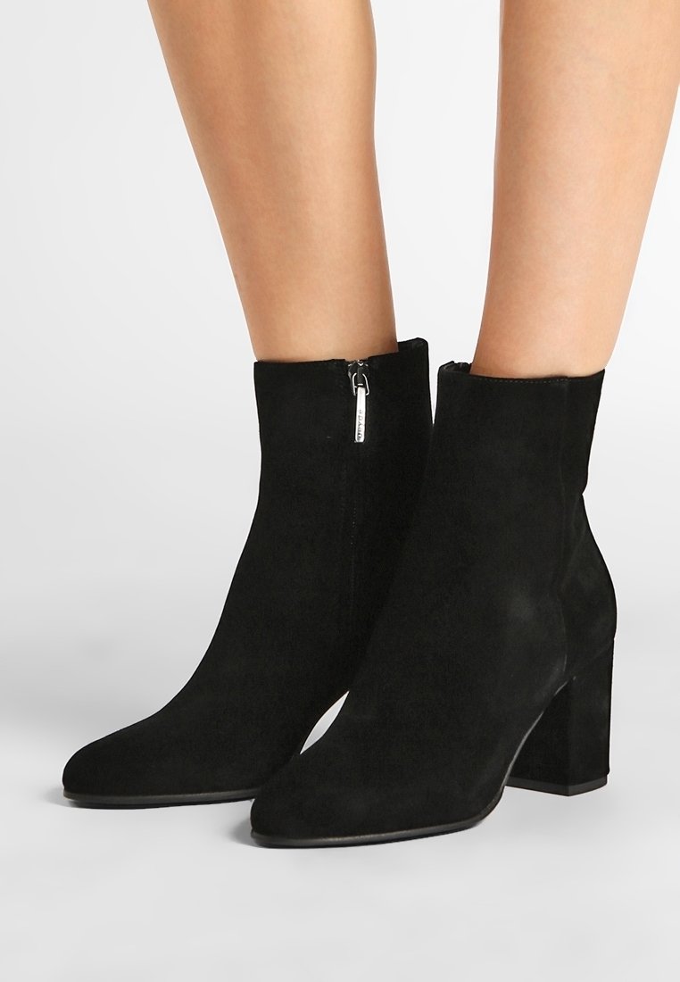 Bottes de cheville en suède noir avec talons carrés et fermetures éclair latérales portées sur jambes nues, présentées sur un fond clair uni.