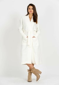 Biały, prążkowany długi kardigan z paskiem i kieszeniami. Model nosi beżowe botki do kostek z bocznym zamkiem. Prosty, teksturowany wzór tkaniny.