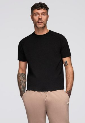 T-shirt noir à manches courtes dans une coupe décontractée, avec un col rond et des manches raglan, associé à un pantalon de jogging beige à cordon de serrage.