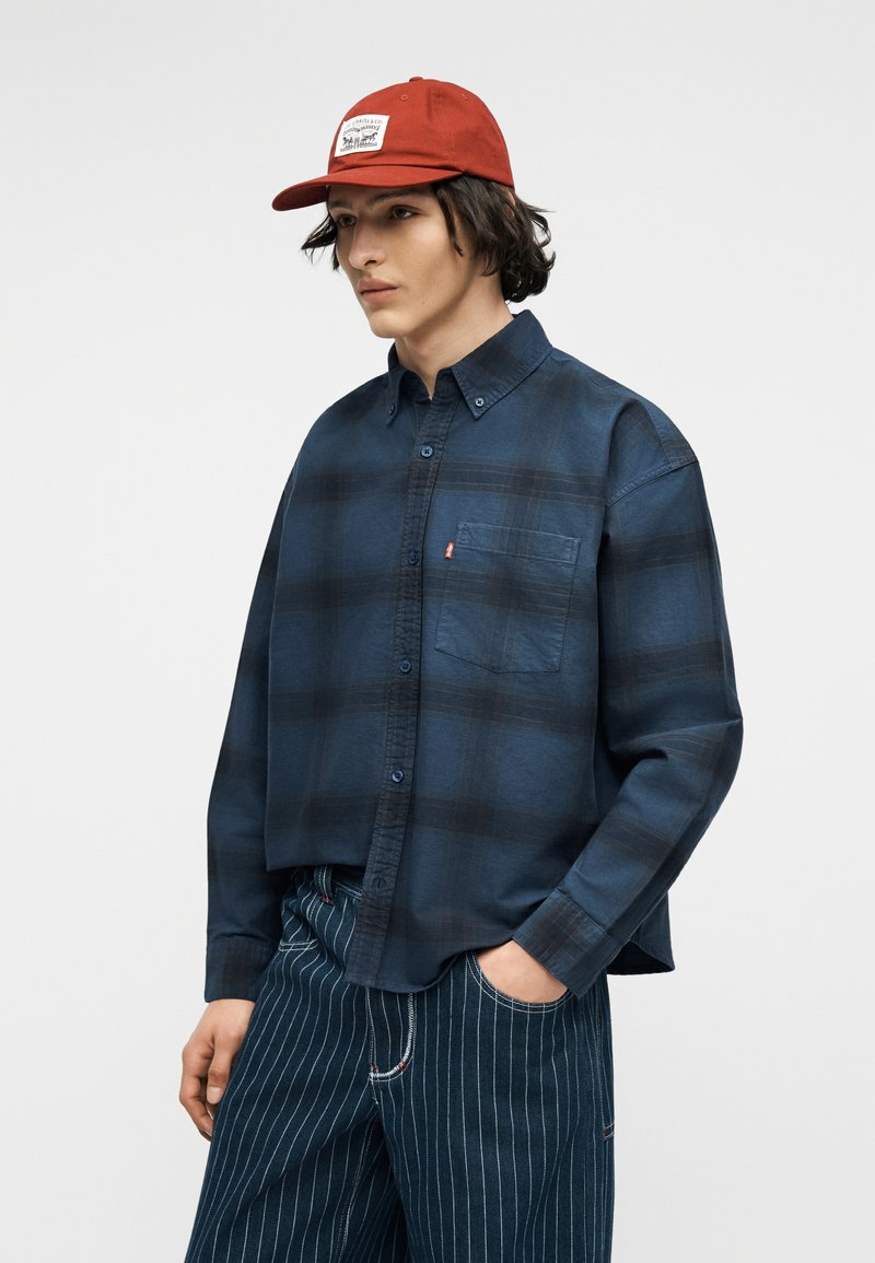 Jeune homme portant une casquette rouge, une chemise à carreaux bleu foncé à boutons, et un pantalon bleu à rayures fines, debout avec une main dans la poche.