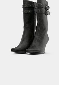 Black wedge-heeled ankle boots with side zippers, two studded straps, and buckles on a white background.