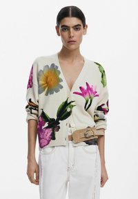 Cardigan fleuri en crème avec des motifs vibrants de violet, jaune et vert. Doté de boutons sur le devant et de poignets côtelés. Porté sur un pantalon blanc.